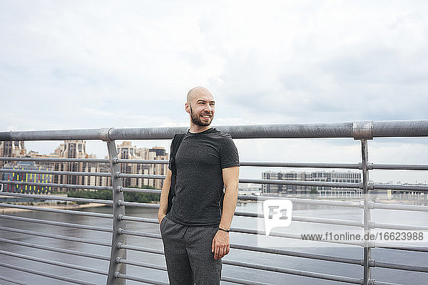 Lächelnder junger Mann  der wegschaut  während er auf einer Brücke in der Stadt am Geländer steht