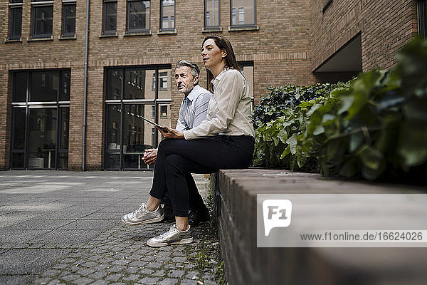 Geschäftsfrau und Mann sitzen auf einer Stützmauer vor einem Gebäude