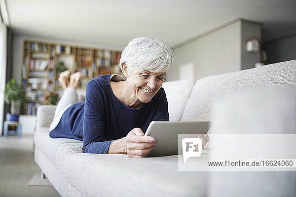Ältere Frau mit digitalem Tablet auf dem Sofa zu Hause liegend