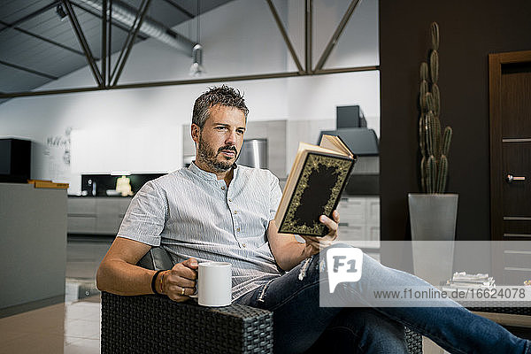 Entspannter männlicher Freiberufler  der mit einer Kaffeetasse auf einem Stuhl im Büro sitzt und ein Buch liest