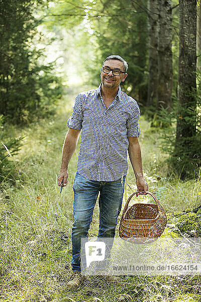 Lächelnder Mann  der einen Korb hält  während er im Wald steht