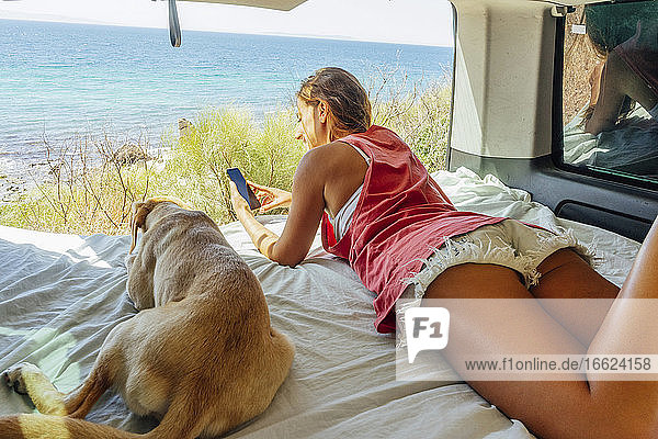 Woman using smart phone while lying with dog on bed in motor home in front of sea