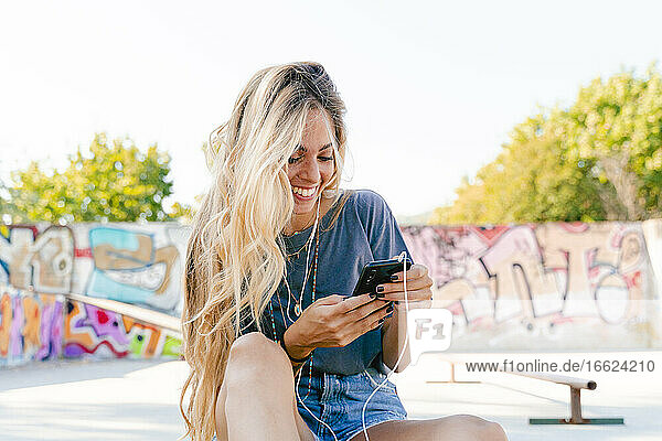 Glückliche junge blonde Frau  die ihr Smartphone benutzt  während sie mit einem Skateboard im Park sitzt