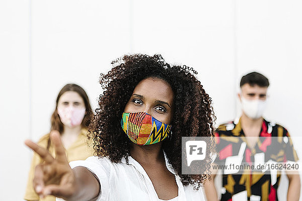 Young woman doing gesture while wearing face mask with friends standing against wall