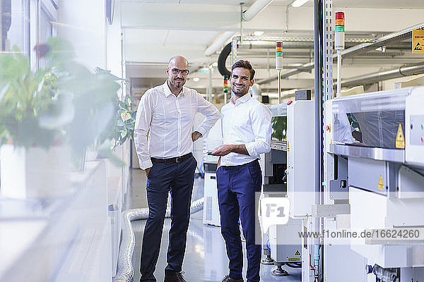 Lächelnde männliche Ingenieure  die eine Fernbedienung in der Hand halten  während sie an einer Maschine in einer Fabrik stehen