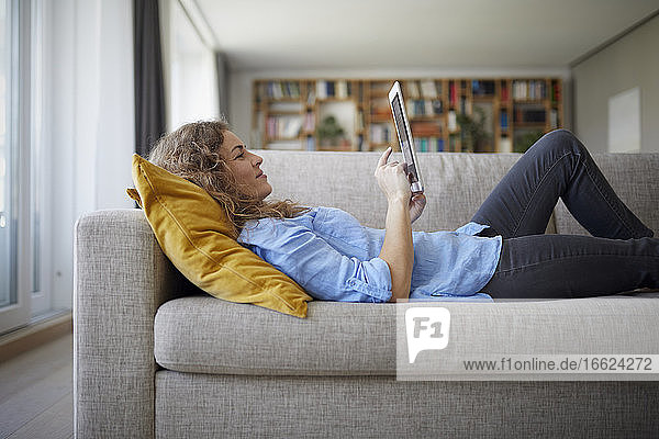 Mittlere erwachsene Frau  die ein digitales Tablet benutzt  während sie zu Hause auf dem Sofa liegt