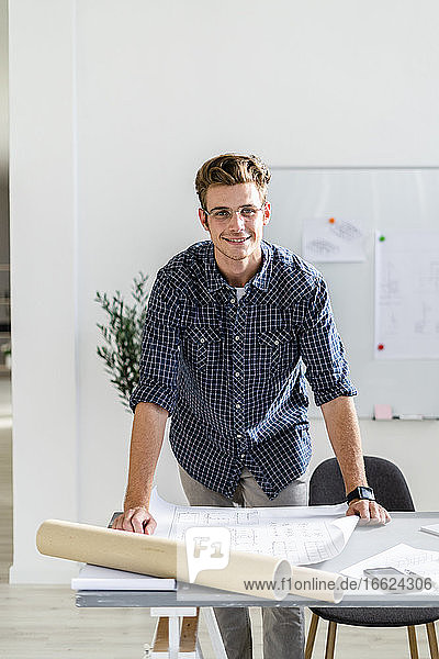 Smiling architect leaning on desk while standing at office