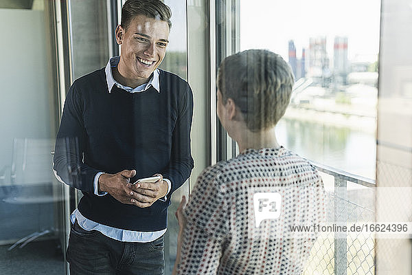 Lächelnder Geschäftsmann mit Smartphone im Gespräch mit einer Mitarbeiterin im Büro durch ein Fenster gesehen