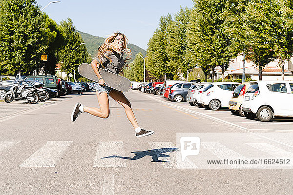 Fröhliche junge Frau  die ein Skateboard hält und über einen Zebrastreifen in der Stadt springt