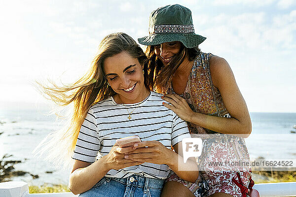 Lächelnde junge Freundinnen benutzen ihr Smartphone am Meer während eines sonnigen Tages