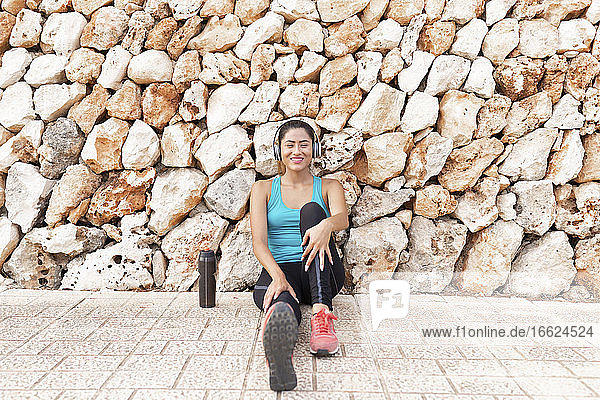Lächelnde schöne junge Frau hört Musik über Kopfhörer  während sie nach dem Sport an einer Steinmauer sitzt