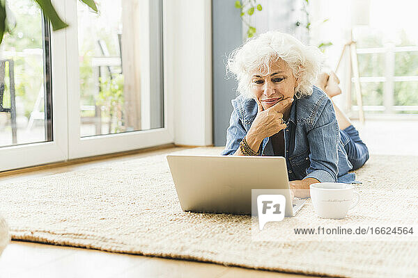 Reife Frau liegt auf der Vorderseite  während sie zu Hause am Laptop arbeitet