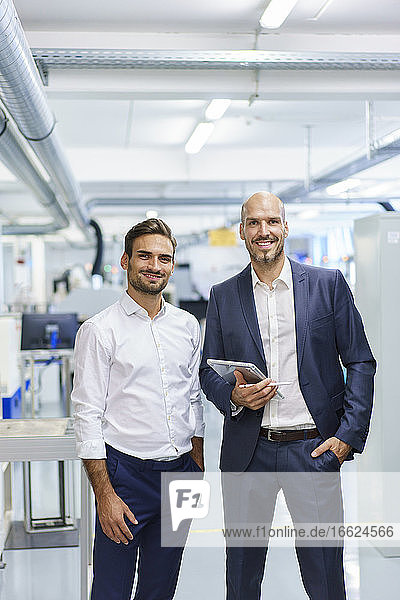 Smiling mature businessman holding digital tablet while standing by young male engineer at factory