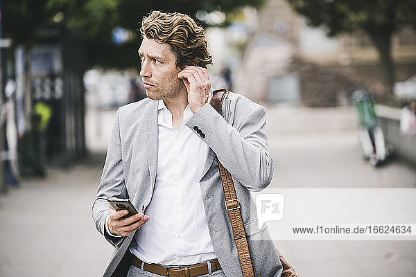 Man adjusting in-ear headphones while standing at sidewalk in city