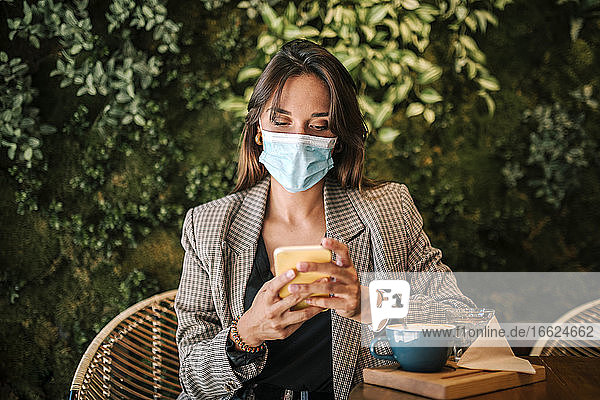 Frau  die eine Textnachricht auf ihrem Smartphone schreibt und dabei eine Gesichtsmaske trägt  während sie in einem Cafe sitzt