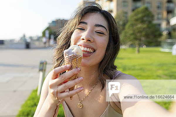Lächelnde Frau isst Eis  während sie ein Selfie in einem öffentlichen Park an einem sonnigen Tag macht
