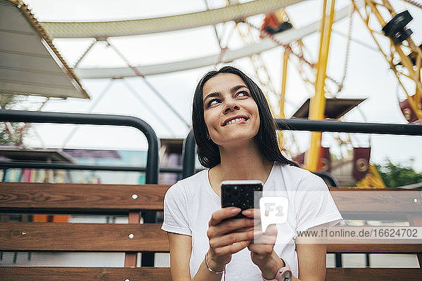 Junge Frau benutzt ihr Smartphone  während sie auf einer Bank im Vergnügungspark sitzt