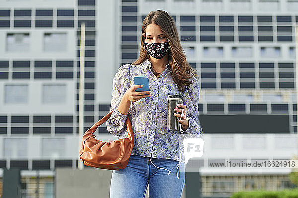 Junge Frau mit Gesichtsmaske  die ein Mobiltelefon benutzt  während sie in der Stadt steht