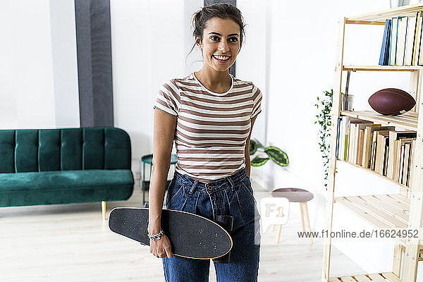 Lächelnde junge Geschäftsfrau  die ein Skateboard in der Hand hält  während sie am Regal eines kreativen Arbeitsplatzes steht