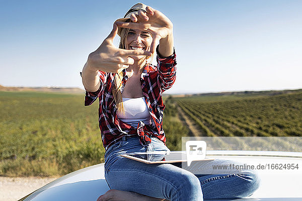 Lächelnde junge Frau zeigt Fingerrahmen  während sie auf dem Autodach sitzt