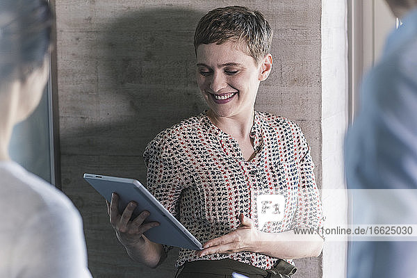 Lächelnde Geschäftsfrau  die ein digitales Tablet hält  während sie sich mit Kollegen im Büro unterhält