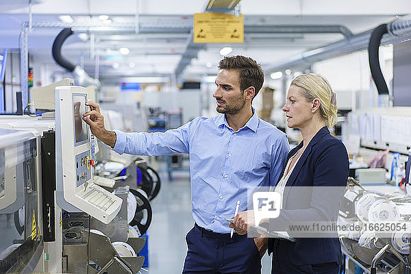 Reife Geschäftsfrau steht neben einem jungen männlichen Techniker  der in einer beleuchteten Fabrik Maschinen bedient
