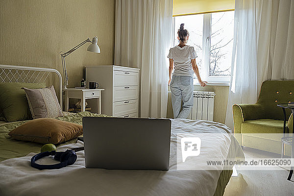 Mid adult woman looking out of window while standing in bedroom
