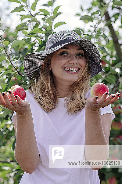 Fröhliche Frau mit Hut und Äpfeln in der Hand auf einem Bauernhof