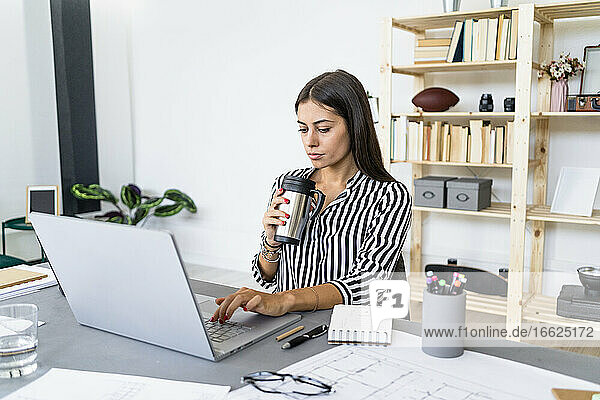 Selbstbewusste schöne Geschäftsfrau  die einen Laptop benutzt und am Schreibtisch im Büro trinkt