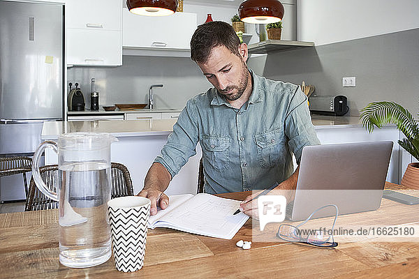 Selbstbewusster männlicher Freiberufler  der mit seinem Laptop am Esstisch sitzt und von zu Hause aus arbeitet und aus einem Buch liest