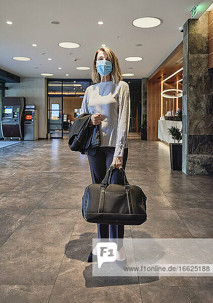 Frau mit Gesichtsmaske  die eine Tasche trägt  steht in einer Hotellobby