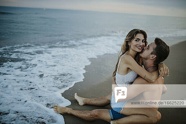 Mann küsst Frau  die auf seinem Schoß am Strand sitzt