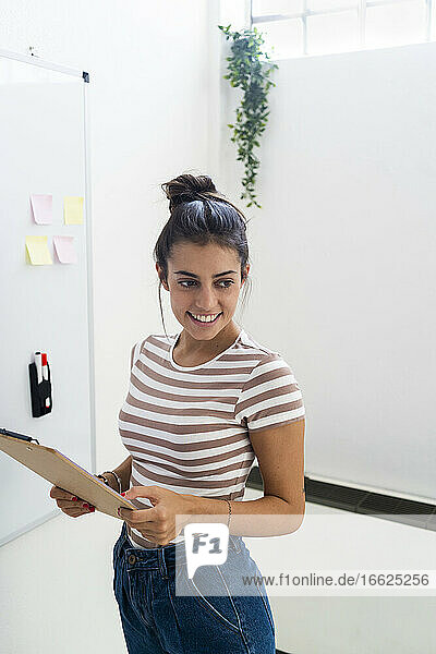 Smiling beautiful female design professional holding clipboard while looking away at creative office