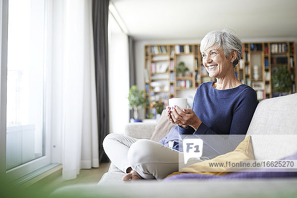 Lächelnde Frau hält eine Kaffeetasse,  während sie zu Hause auf dem Sofa sitzt