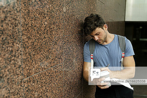 Gut aussehender junger Mann  der in ein Buch schreibt  während er eine Kaffeetasse hält und sich an eine braune Kachelwand in einer U-Bahn-Station lehnt