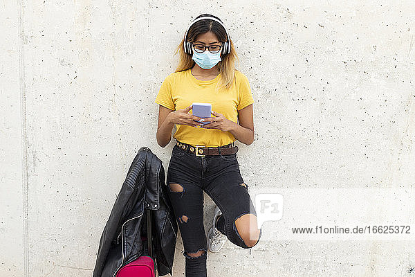 Junge Frau mit Schutzmaske bei der Benutzung eines Smartphones mit Gepäck an einer weißen Wand an einer Bushaltestelle