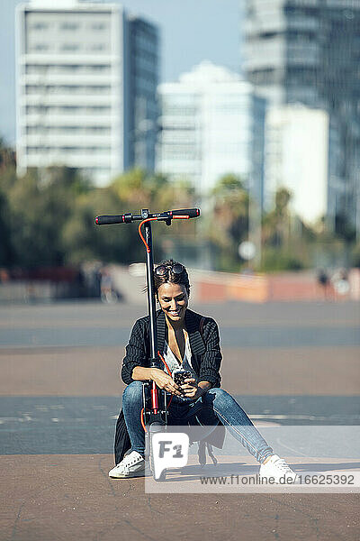 Junge Frau  die ein Mobiltelefon benutzt  während sie auf einem Motorroller in der Stadt sitzt