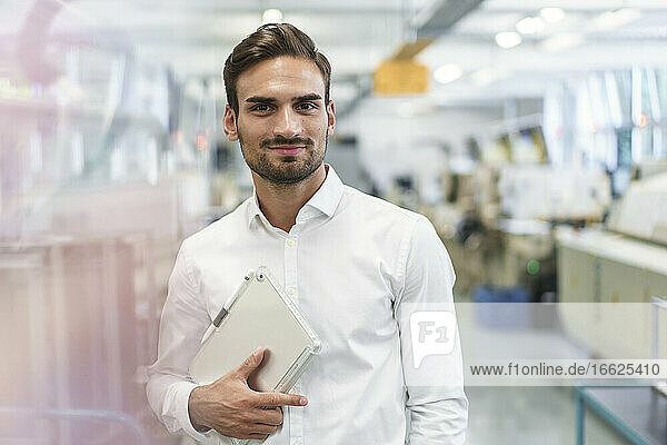 Selbstbewusster junger männlicher Ingenieur  der ein digitales Tablet hält  während er in einer beleuchteten Fabrik steht