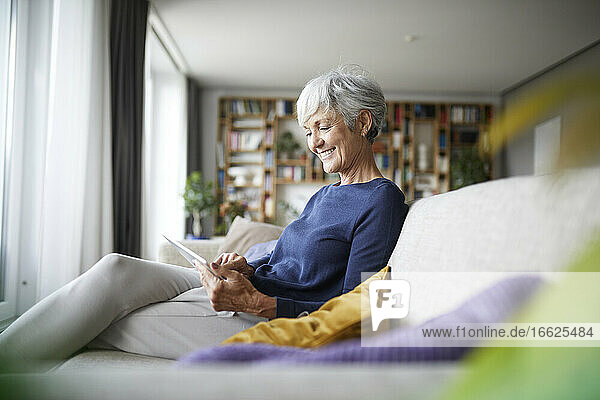 Lächelnde Frau  die in ein digitales Tablet schaut  während sie zu Hause auf dem Sofa sitzt