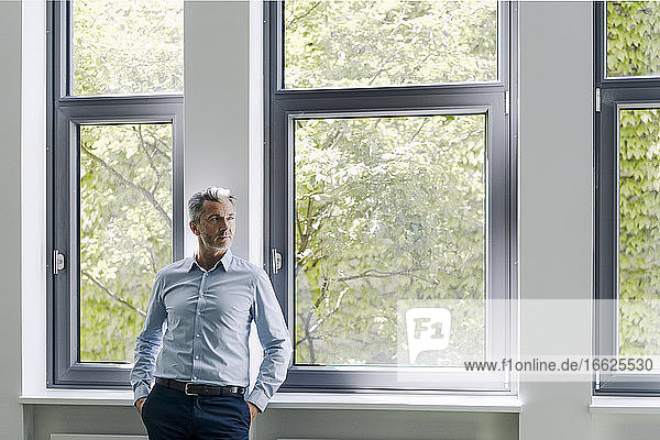 Businessman looking away while leaning by window at office
