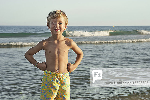 Smiling boy with hand on hip standing against sea at beach