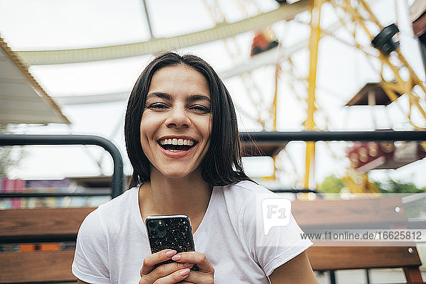 Frau hält Smartphone lachend auf einer Bank im Vergnügungspark sitzend