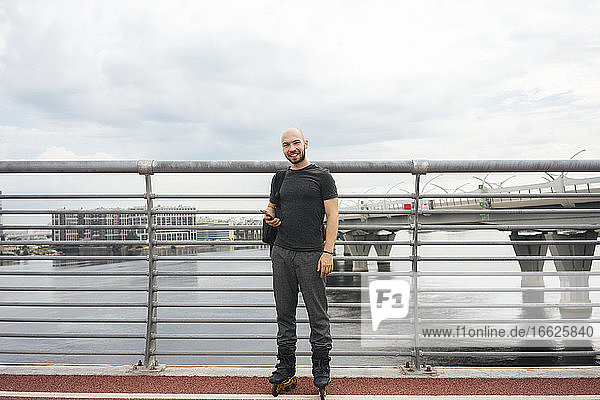 Lächelnder junger Mann mit Inline-Skates  der auf einer Brücke stehend sein Smartphone benutzt