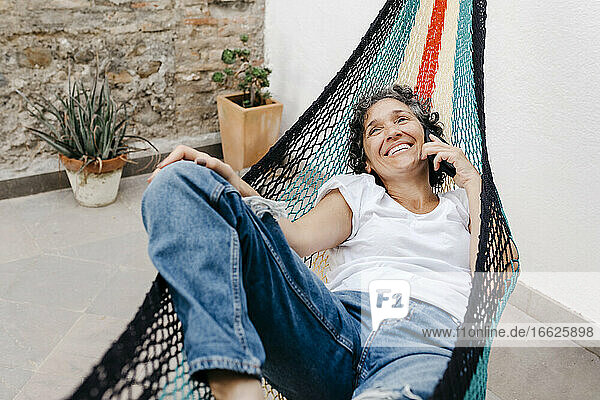Lächelnde Frau  die über ihr Smartphone spricht  während sie sich in einer Hängematte im Garten entspannt