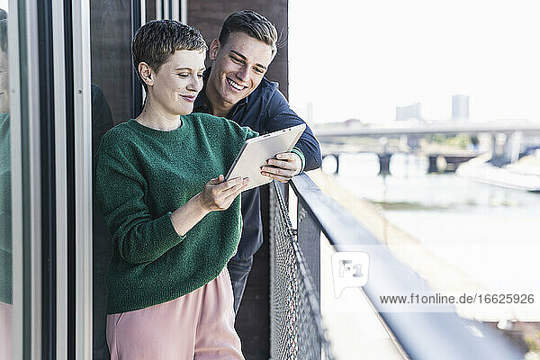 Lächelnde Kollegen diskutieren über ein digitales Tablet  während sie auf einem Balkon im Büro stehen