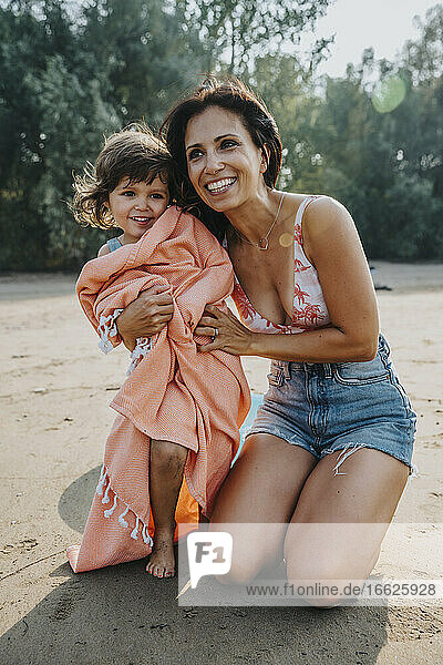 Lächelnde Mutter schaut weg  während sie ihrer Tochter am Strand ein Handtuch umlegt