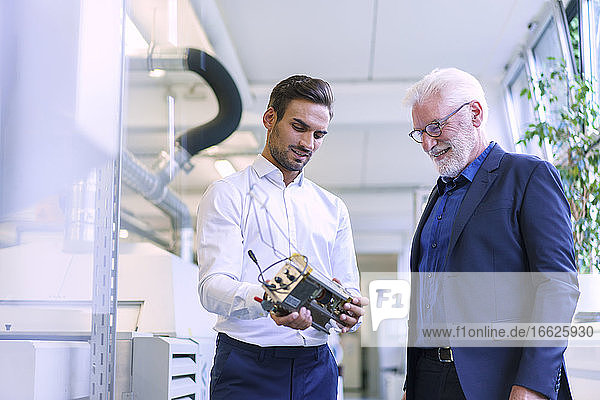 Junger männlicher Ingenieur  der ein Maschinenteil hält  während er mit einem lächelnden Senior Manager in einer Fabrik diskutiert