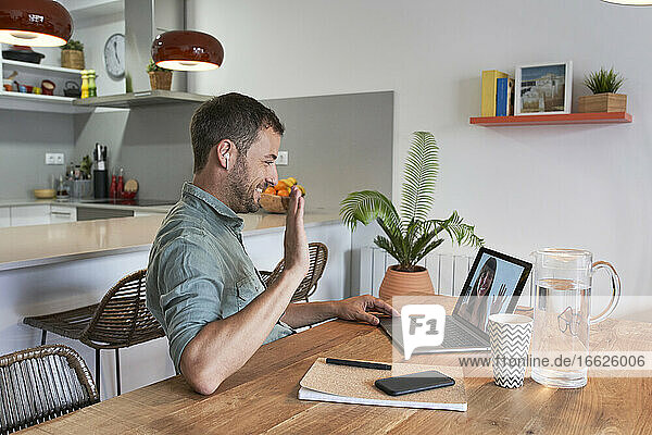 Lächelnder Geschäftsmann  der einer Kollegin per Videoanruf auf dem Laptop zuwinkt  während er von zu Hause aus arbeitet