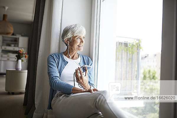 Aktive ältere Frau  die Musik auf ihrem Smartphone hört und aus dem Fenster schaut  während sie zu Hause sitzt