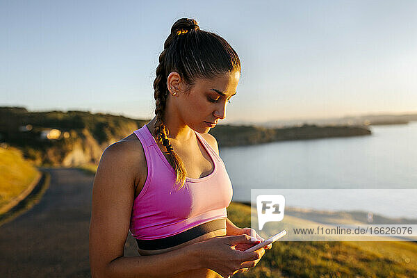 Junge Frau  die ihr Smartphone benutzt  während sie auf einer Klippe gegen den klaren Himmel bei Sonnenuntergang steht